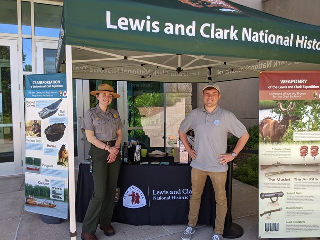 Two rangers stand in front of outdoor visitor station. Tent reads Lewis and Clark Trail. Portable history displays.