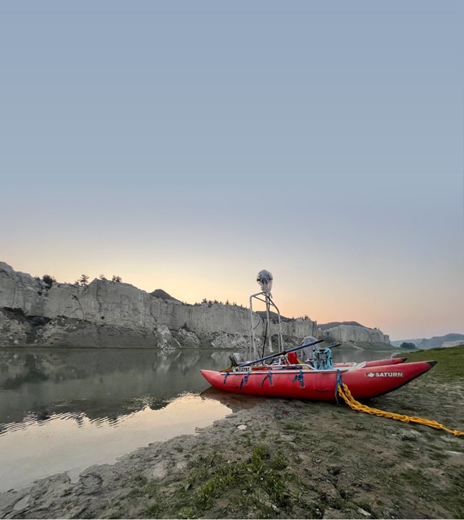 Red double hull raft sits on riverbank. Camera mounted up high. White cliffs and sunset beyond.