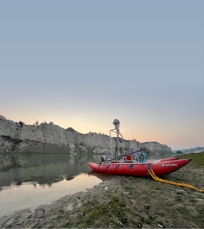 Terrain 360 Upper Missouri Breaks Red double hull raft sits on riverbank. Camera mounted up high. White cliffs and sunset beyond.