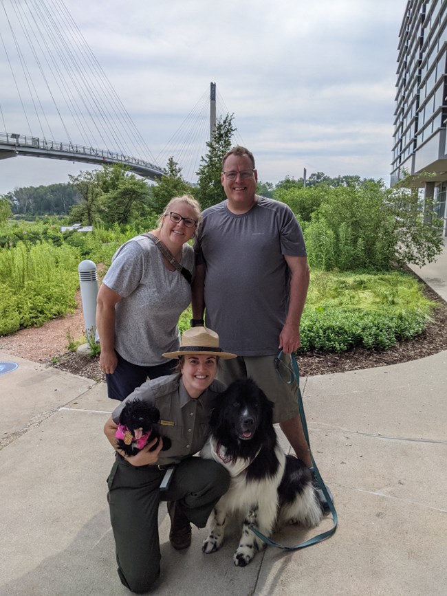 VCO Ranger shannon with Newfoundland dog and visitors_2021 Two visitors and their dog pose with ranger at urban park. Dog is a black Newfoundland. Ranger holds black dog stuffed toy.