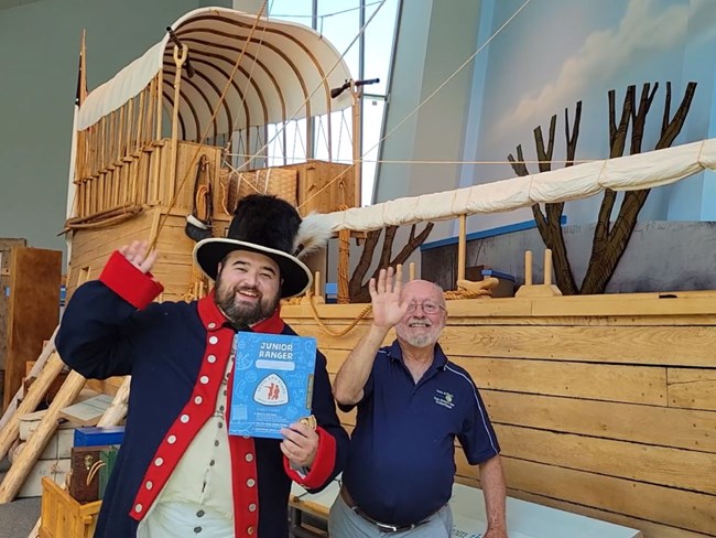 Two men, one a reenactor in early 1800s US military dress wave. Hold blue junior ranger book. Keelboat display behind.