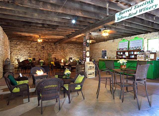A tasting room with tables and chairs is lined with a brick wall and wooden beams.