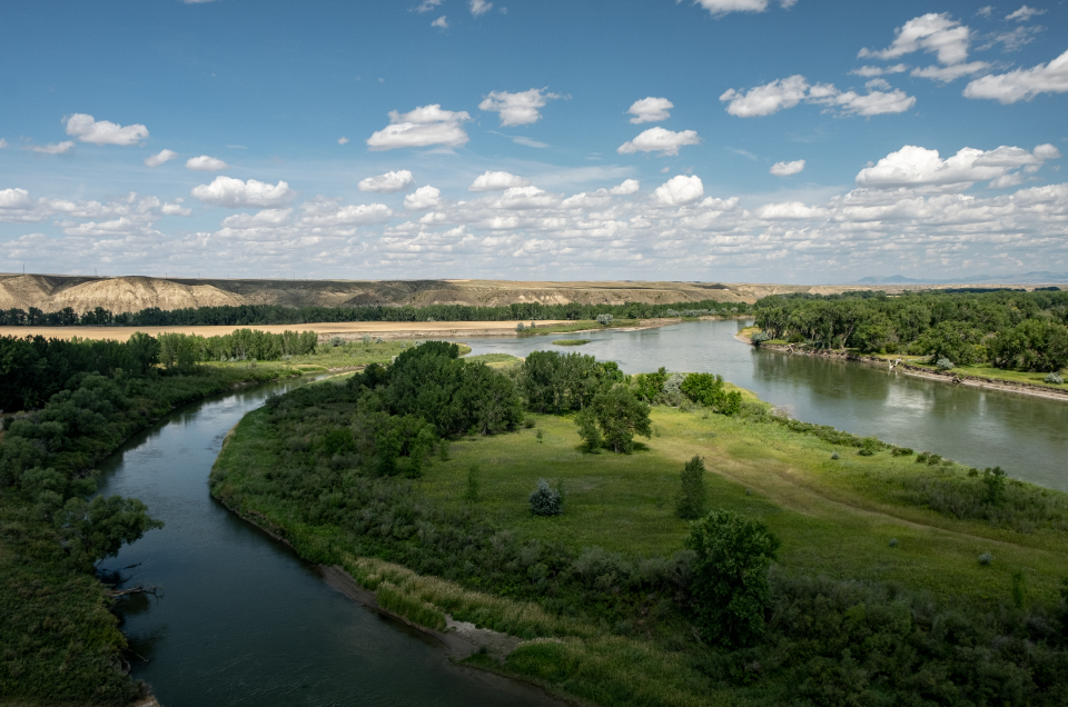 The forks of two rivers meet under a blue sky with mountains behind.