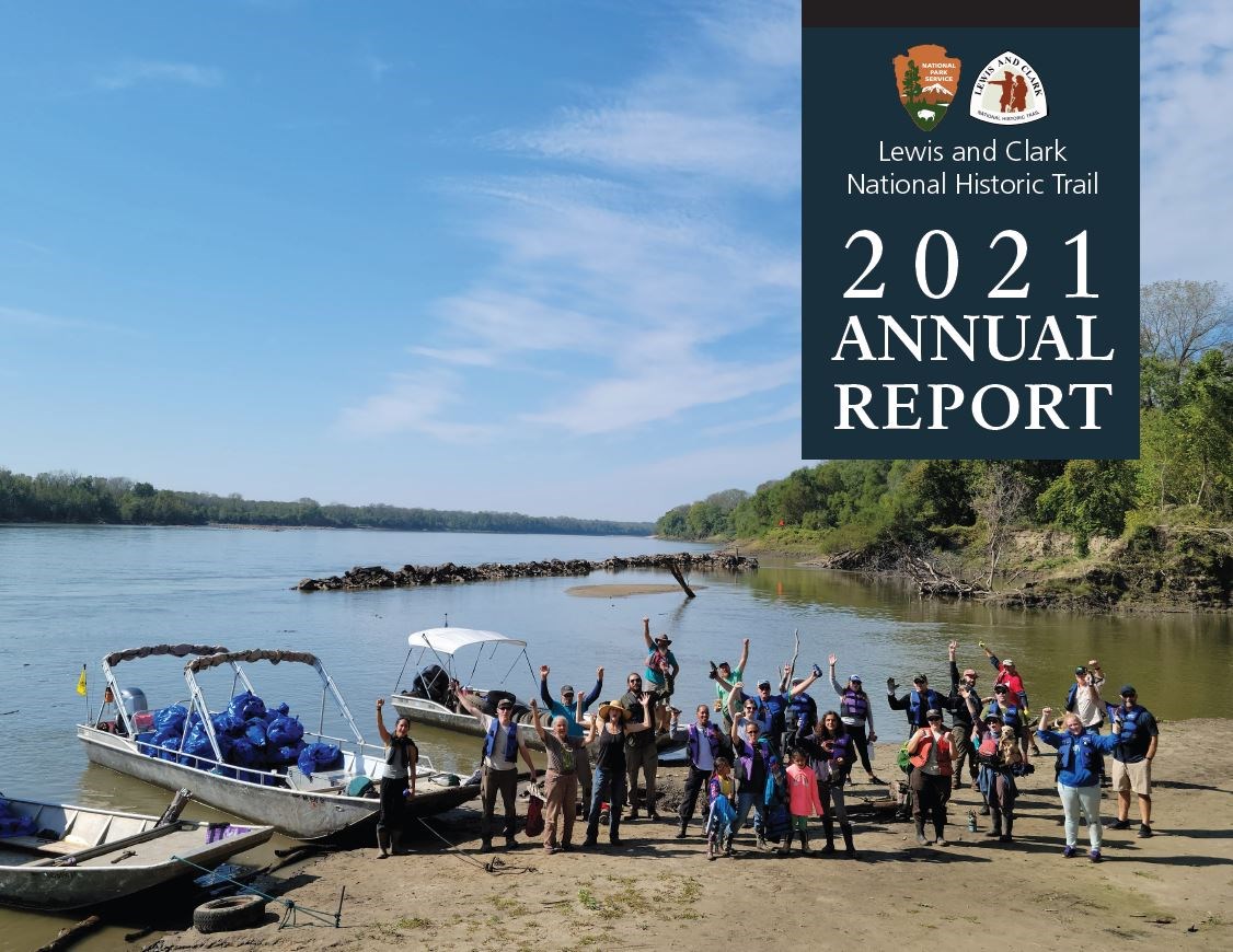 Lewis and Clark National Historic Trail Annual Report 2021. Lewis and Clark Trail Logo. National Park Service Logo. 25 people stand on riverbank and wave. 3 boats behind. Leafy scrubby trees line shores.