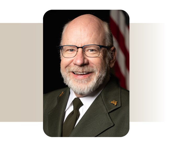 Man in Park Service Class A Uniform Smiles. He wears glasses and has a white beard.