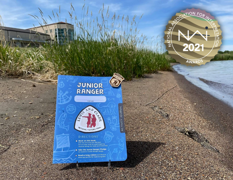 A blue activity booklet and wooden badge are displayed along a lake shore with a visitor center behind. Booklet features a Lewis and Clark Trail logo and reads: Junior Ranger.