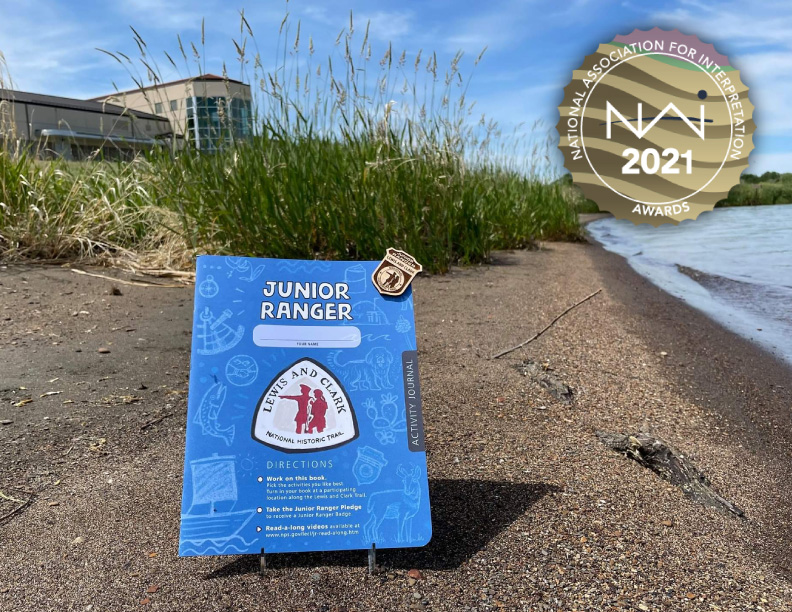 NAI-media-award-2021-Lewis and Clark Trail Junior Ranger A blue activity booklet and wooden badge are displayed along a lake shore with a visitor center behind. Booklet features a Lewis and Clark Trail logo and reads: Junior Ranger.