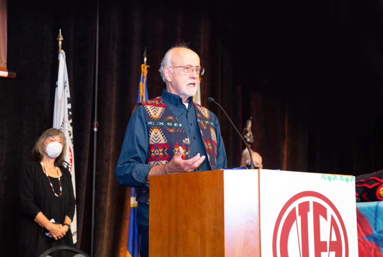 Man at podium addresses audience. He wears a vest in a tribal pattern.