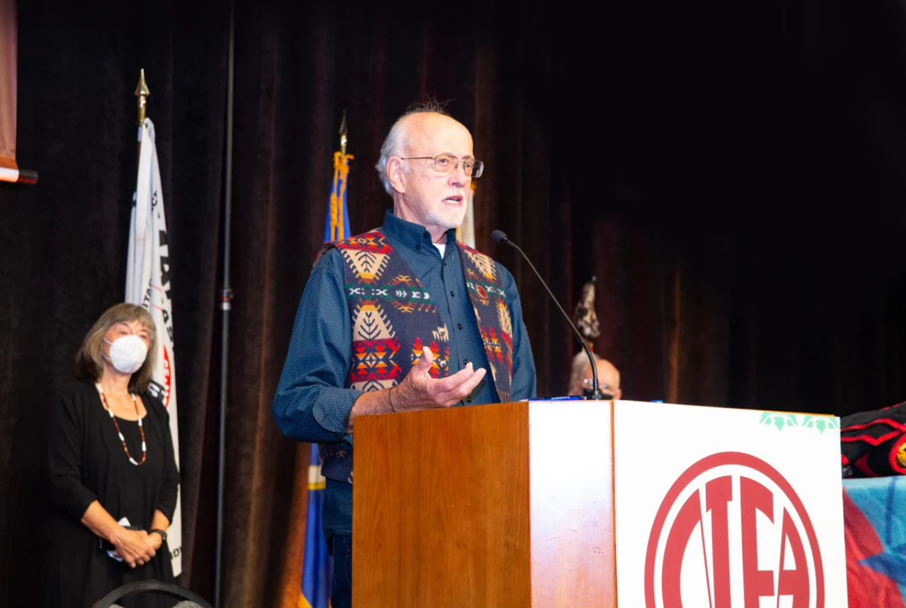 NIEA HTL Dick Basch at Podium_2021 Man at podium addresses audience. He wears a vest in a tribal pattern.