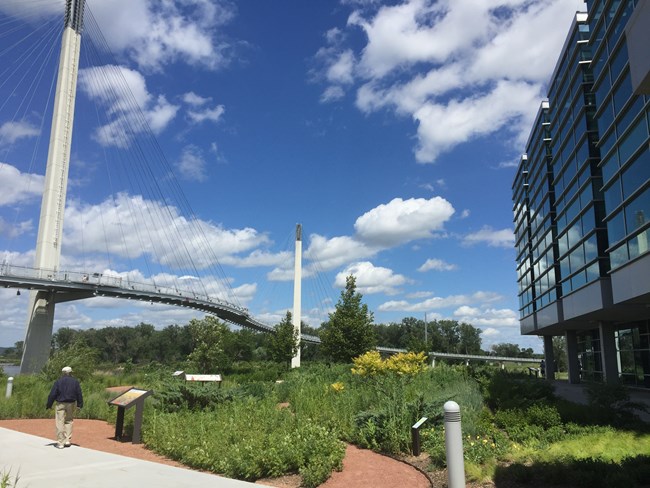 A footbridge spanning the Missouri River with garden, office building, and a person walking