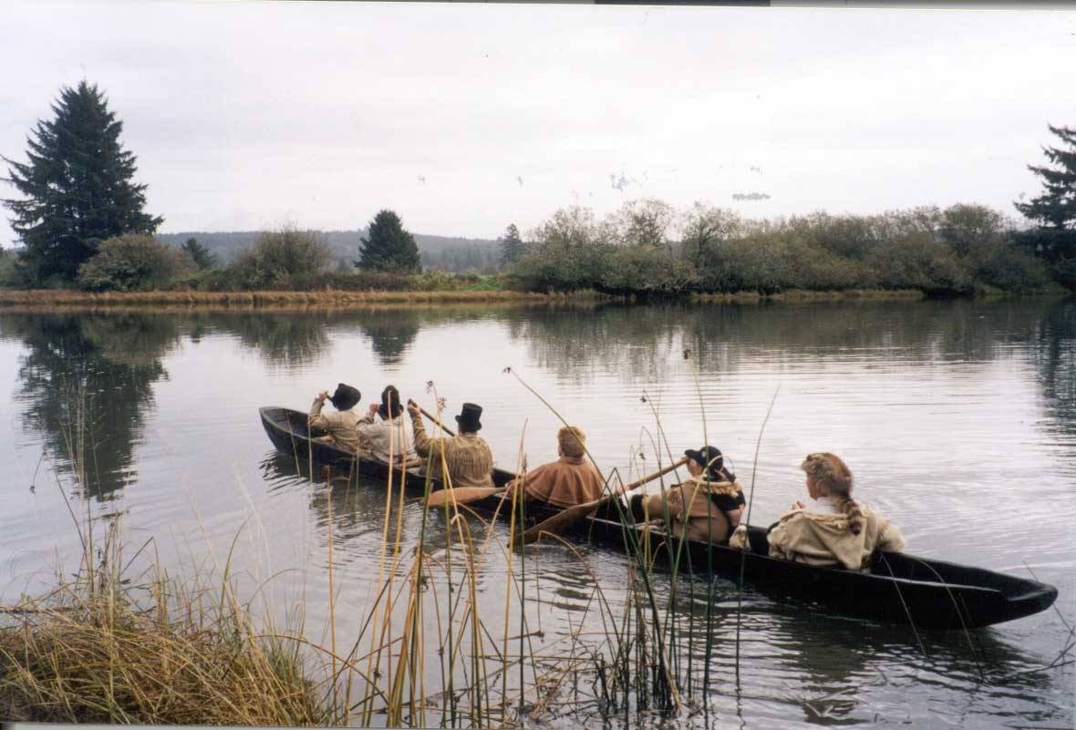 Photo of six people in historic dress paddle a dugout canoe away from a marshy bank.