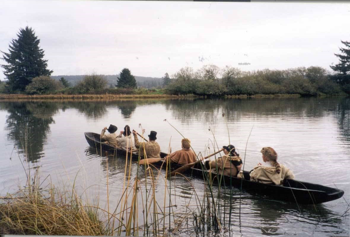 Canoe Paddling at Netul Landing Photo of six people in historic dress paddle a dugout canoe away from a marshy bank.