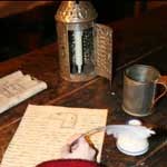 Hand writing on paper with quill pen on wooden desk. Also on the desk is a lantern, tin cup and inkwell.