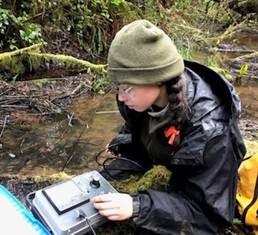 A ranger kneels next to a creek in rain gear while holding a rectangular monitoring tool