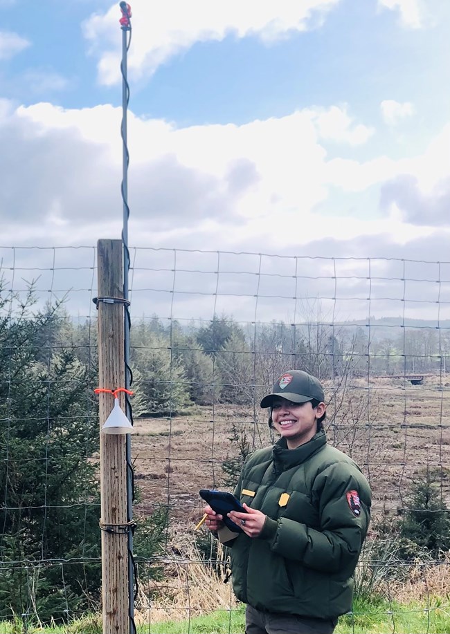 A park ranger in a green uniform and ball cap stands next to a bat recorder attached to a fence post while holding an iPad and smiling at the camera.