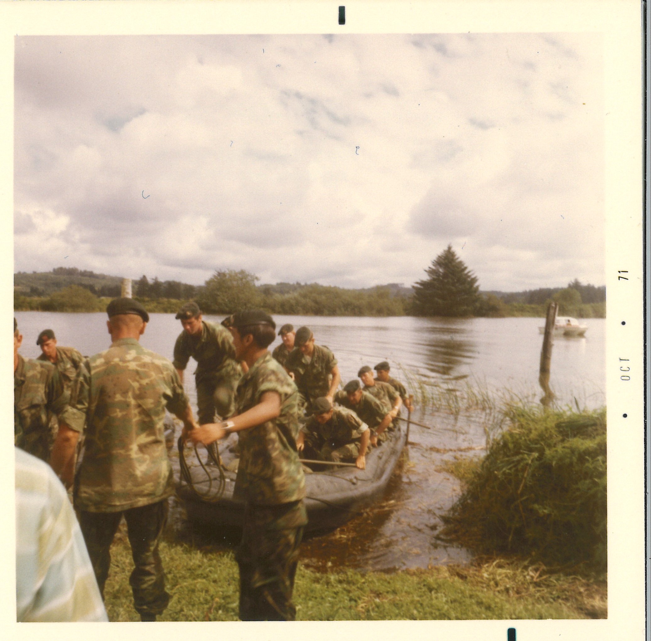 Green berets landing at Fort Clatsop photo 1971 A square color photograph from the 70's. At least 12 men in military uniform with green berets sit on a black raft or stand on the shore holding onto the raft. The raft is floating on a river, with evergreen trees and short shrubs lining the bank.