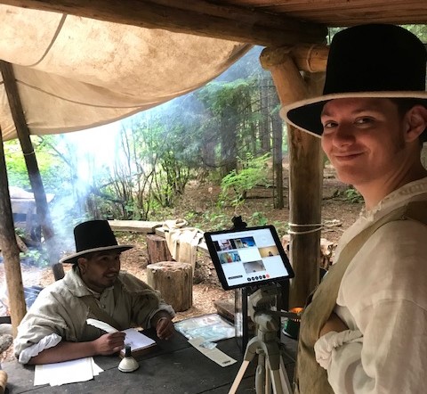 Virtual quill writing demonstration A man wearing a black felt hat, white linen shirt and buckskin vest looks at the camera while using an iPad to livestream another costumed ranger in similar clothes as they demonstrate how to write with a quill pen.