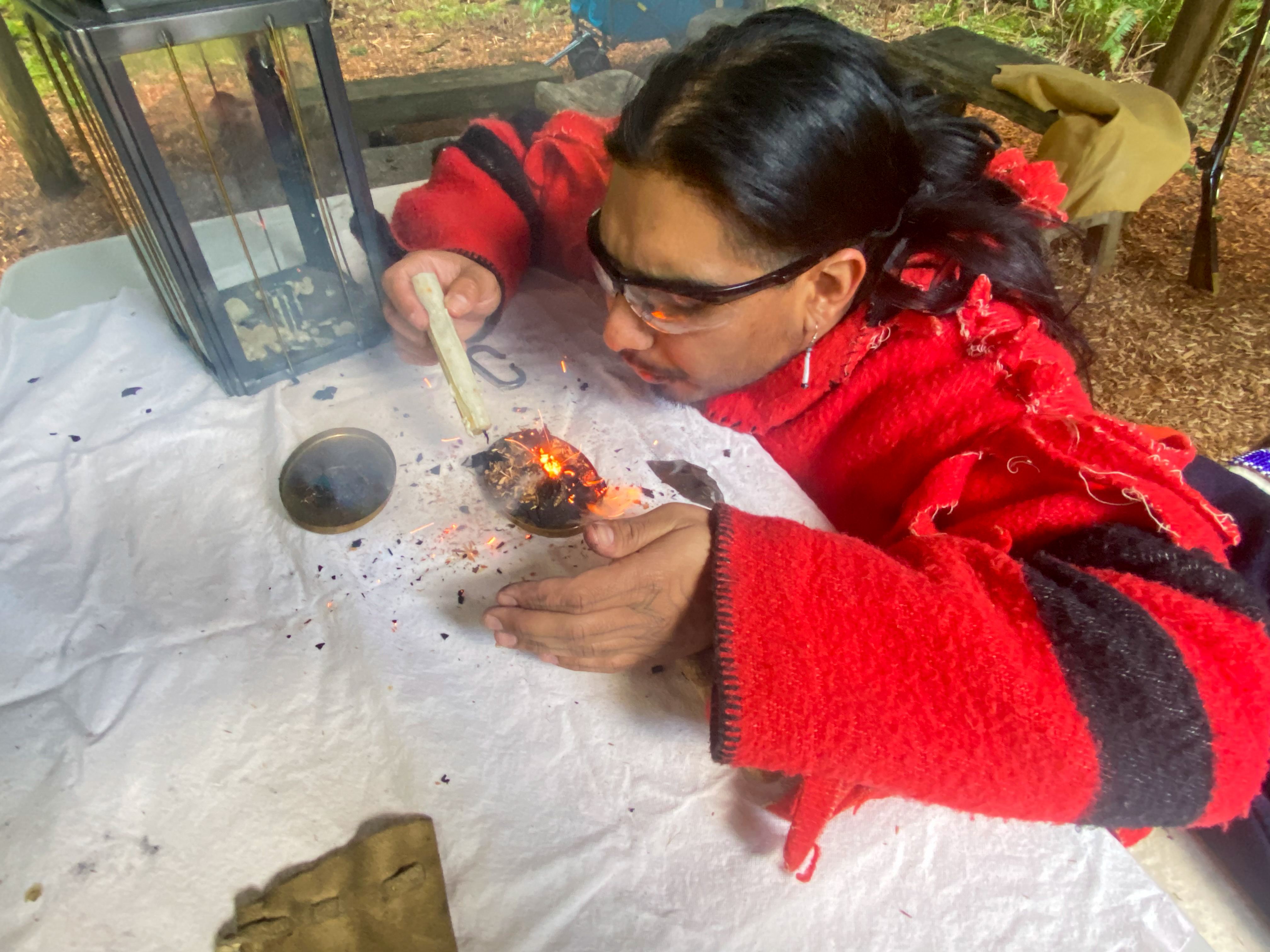 Fire Starting A man wrapped in a red wool blanket coat kneels next to a table, and blows on a spark on a small pile of woodchips to start a fire.