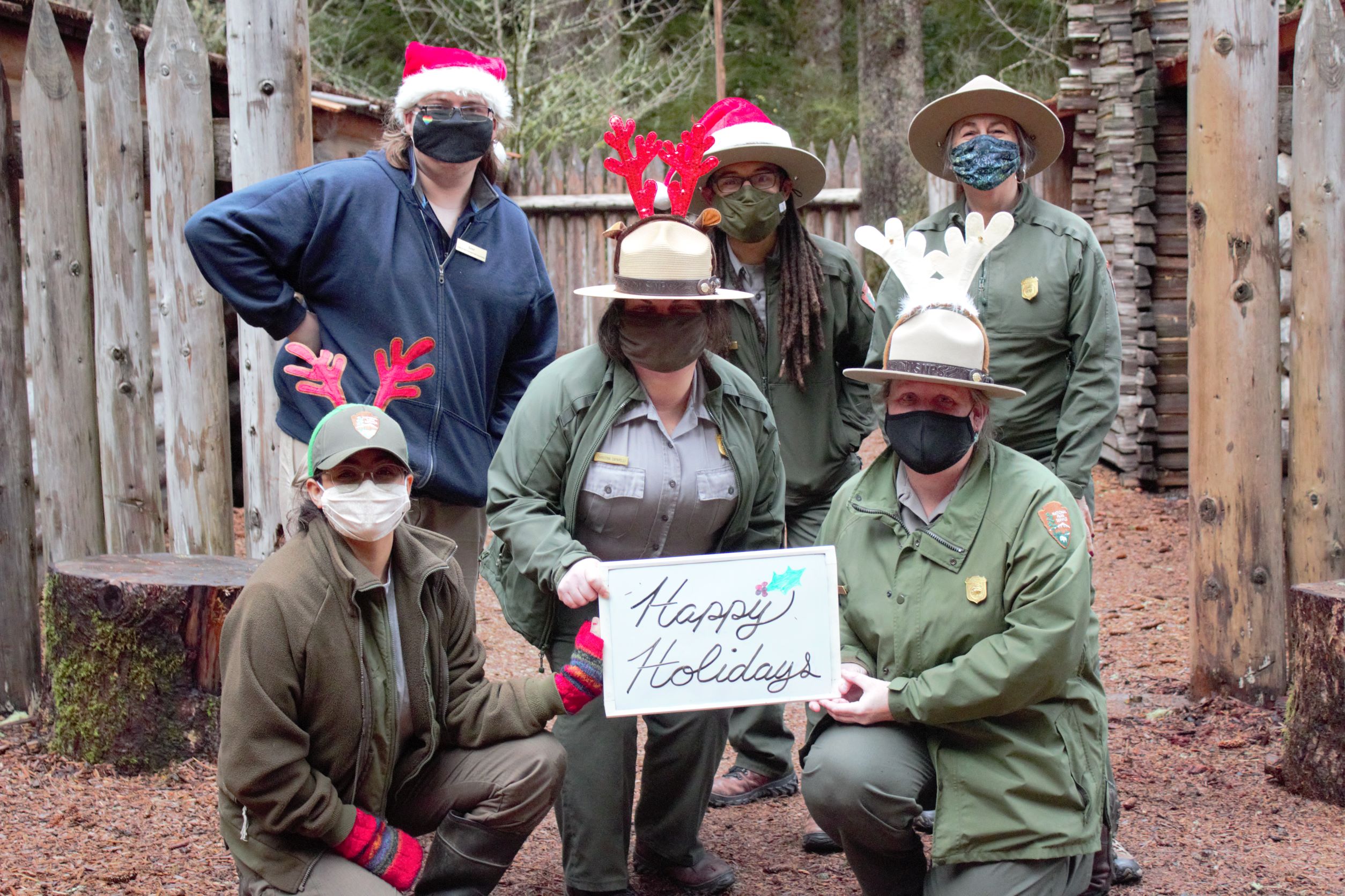 Holiday Photo 5 rangers and one association employee stand together in front of Fort Clatsop holding a sign that says "Happy Holidays." The rangers all wear green uniforms with wide brim hats. On top of the hats are antlers, and red caps with white trim.