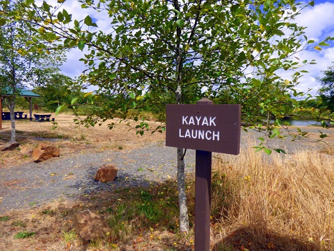 Open area with a gravel trail leading to the river with a sign that says kayak launch