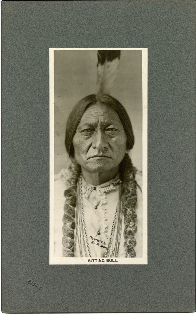 This image depicts a portrait of Sitting Bull, Lakota Chief, in bust view. His hair is braided and wrapped in fur. There is one feather in his hair. He is wearing a leather shirt and several chain necklaces.