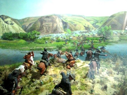 A painting  showing a number of military men on horses crossing a river.  On the other side of the river are high bluffs.