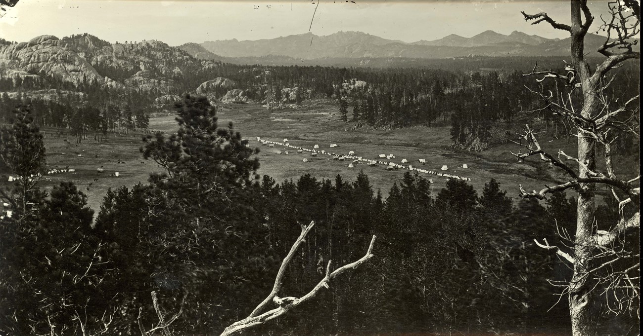 Black and white photograph of a military camp.  The foreground is a heavy vegetated area with the camp in the mid ground with white tents.  The background shows more trees and mountains.