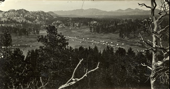 Black and white photograph of a military camp.  The foreground is a heavy vegetated area with the camp in the mid ground with white tents.  The background shows more trees and mountains.
