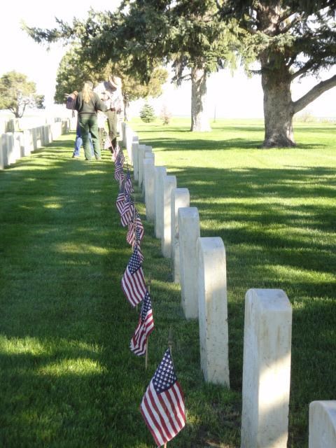 American flags being placed in front of headstones in the Custer National Cemetery for Memorial Day Observances. Miniature american flags placed in front of white headstones in cemetery.