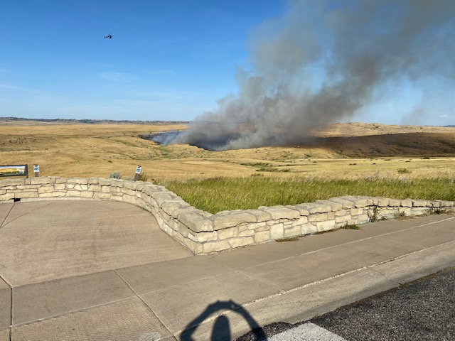 Wildland Fire 8/10/22 Black smoke goes upwards to the sky in a small patch of grassland surrounded by brown and green grass. There is a stone wall about 3 feet high where there are 3 interpretive signs. The sky is bright blue and there are tree spotted hills far away.