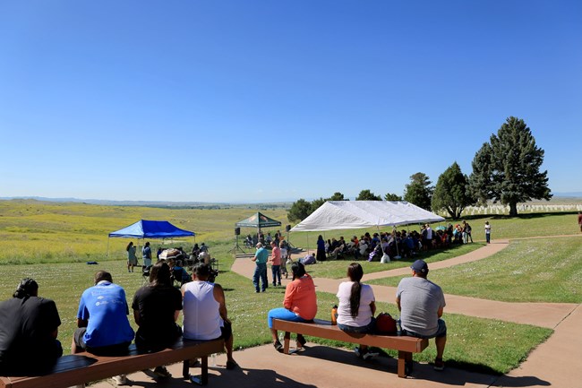 Crowds enjoy the opening ceremonies and speaker series during the 2024 Anniversary of the Battle of Little Bighorn under the shade of tents near the Custer National Cemetery overlooking the main battlefield area.
