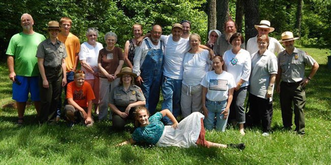 Large group of people gathered in the grass, trees in background. Some wearing pioneer clothing, two in park ranger uniforms, others in regular clothing posing facing forward.