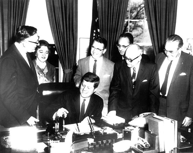 President John Kennedy sitting at desk holding a pen, a piece of paper is on the desk in front of him, while five men and one woman watch.