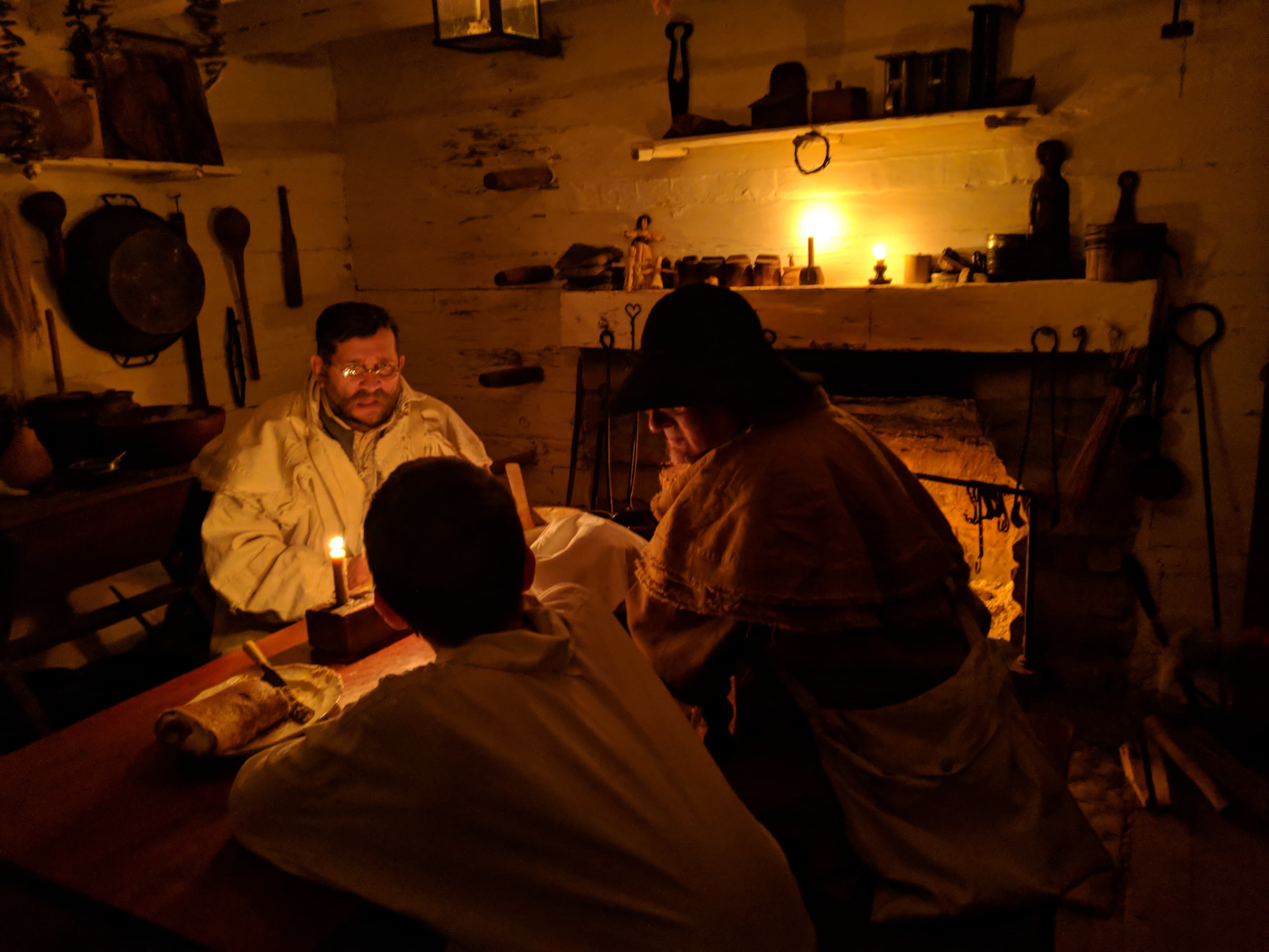 inside candlelit log cabin three people dressed in pioneer clothing gather around a table by the fireplace inside candlelit cabin three people dressed in pioneer clothing gather around a table by the fireplace