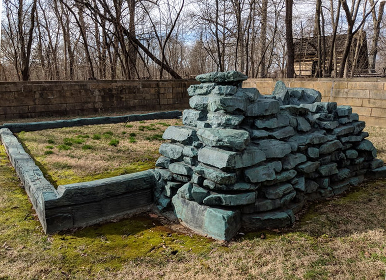 Cabin site memorial and Log cabin Cabin site memorial showing outline of bottom logs and fireplace in foreground, low stone wall from left to right behind memorial, trees bare of leaves and Log Cabin in background.