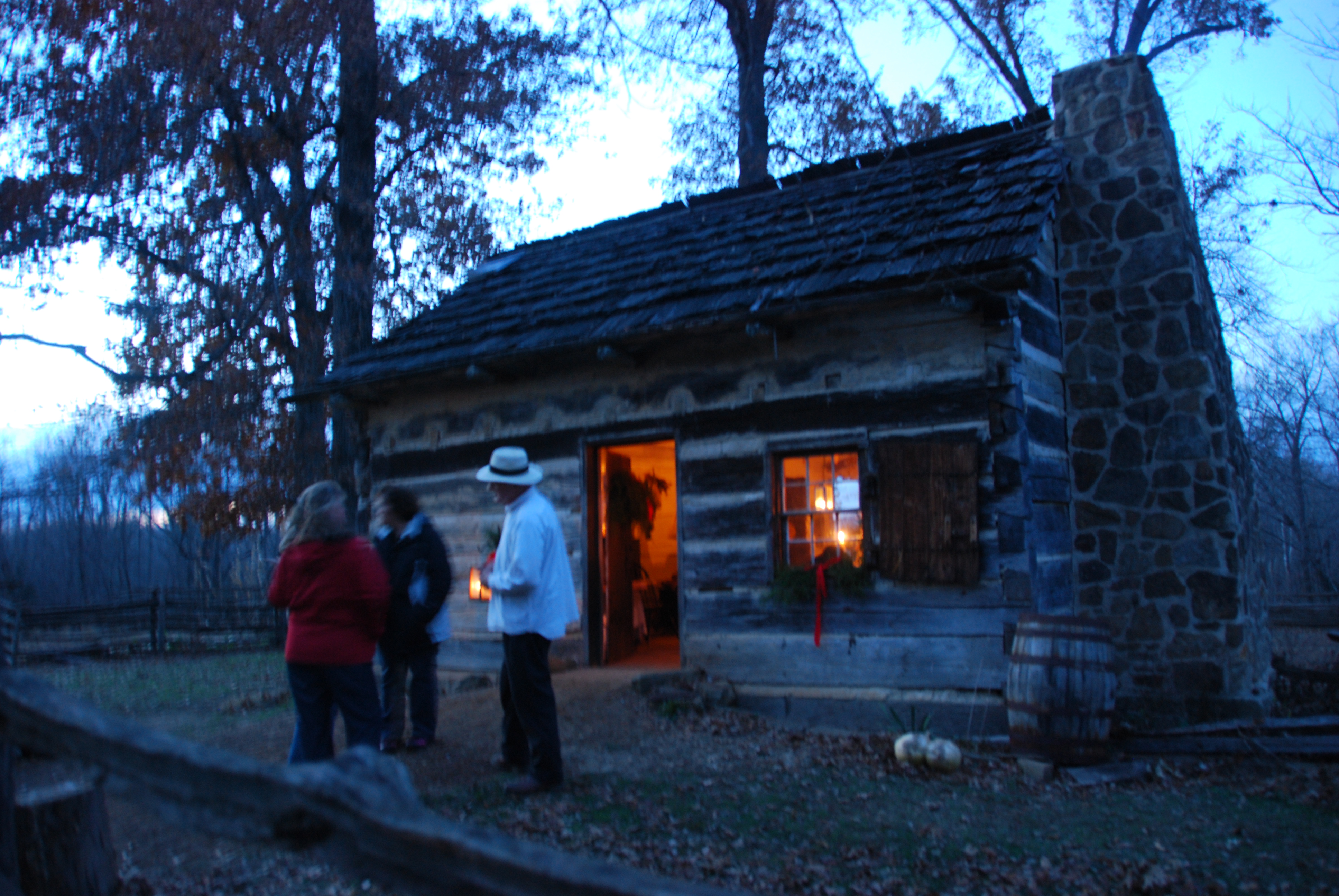 three people standing outside log cabin at dusk, light inside the cabin from candles