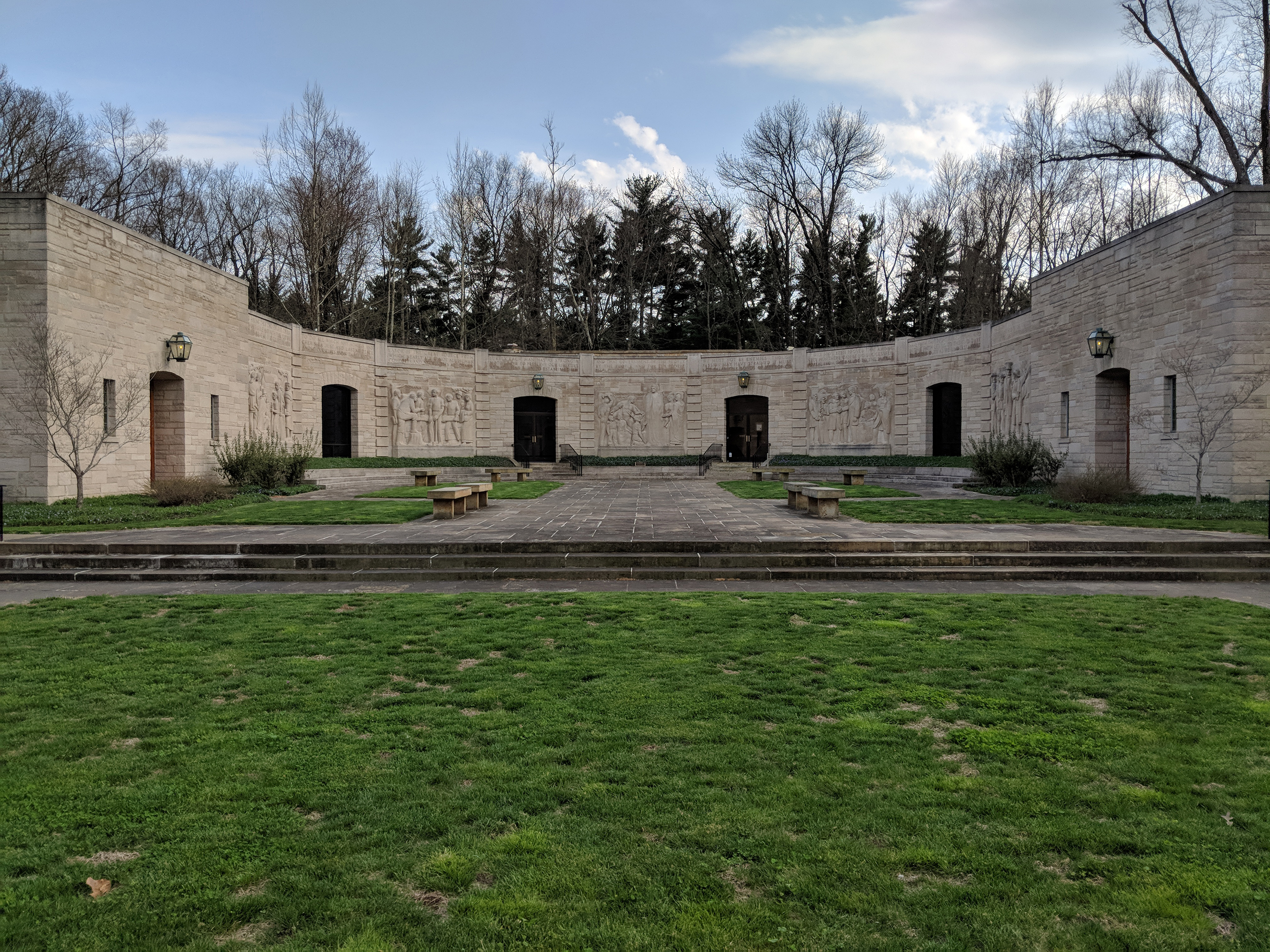 Memorial Visitor Center Curved limestone building with 5 sculptured panels with scenes of people, stone walkway leads to stairs and doorway, grass between walkways, stone benches