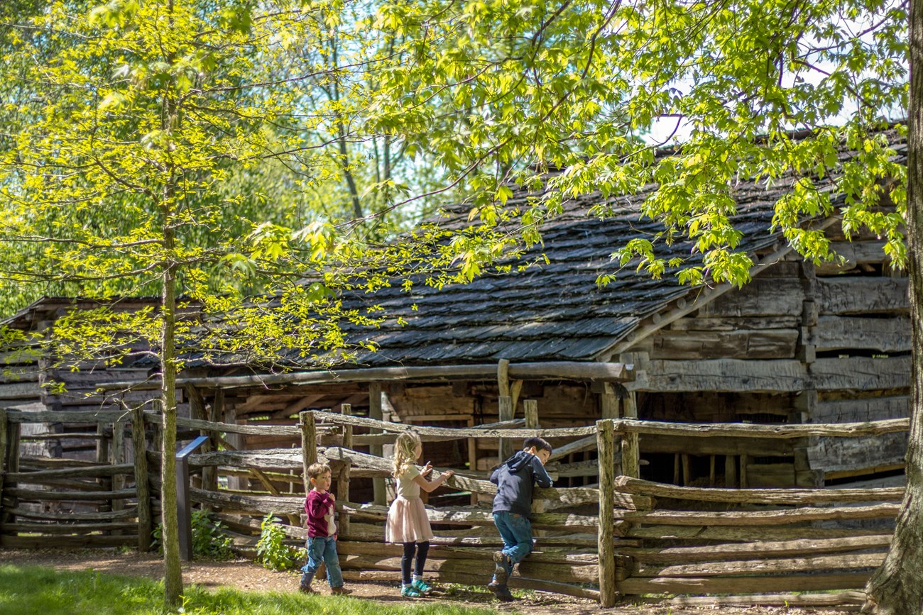 Three young kids at split-rail fence in front of log barn.