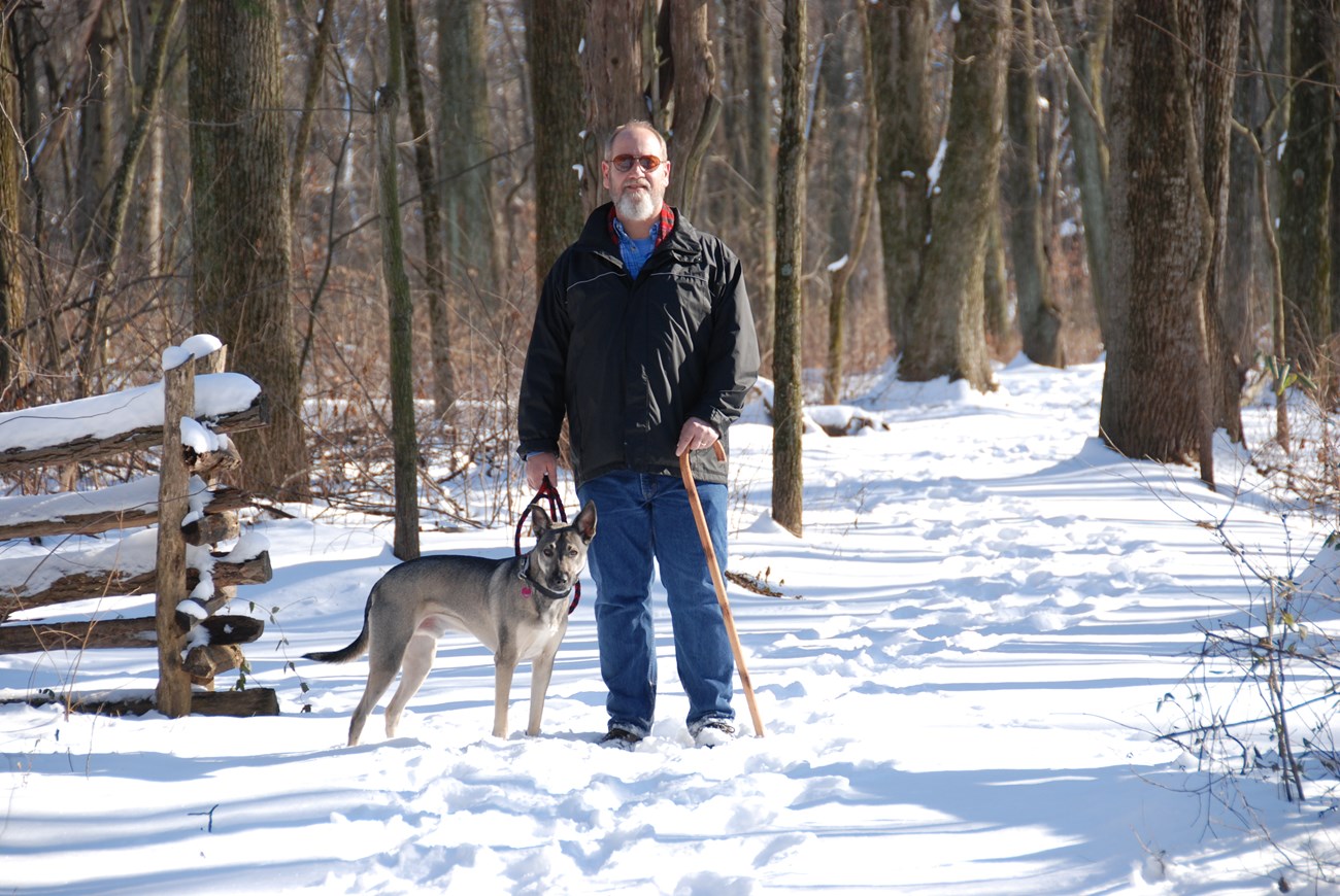 Medium size dog, silver and black, standing on snow covered trail on a leash held by owner in black jacket and jeans. Both looking forward.