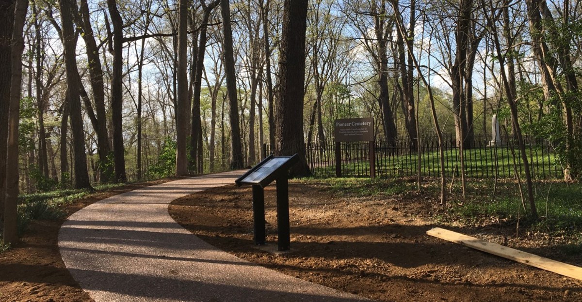 Paved trail winds through woods to Pioneer Cemetery lined by iron fence.