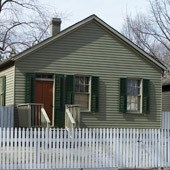 One story pale green house with a small yard and white picket fence