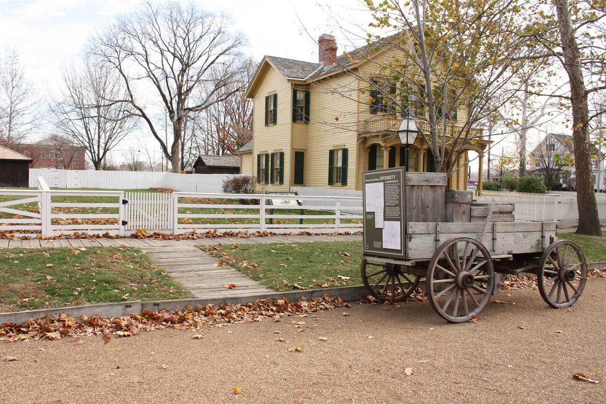 Empty grass lot surrounded by white wood fence with recreated antique wagon stationed on street out front