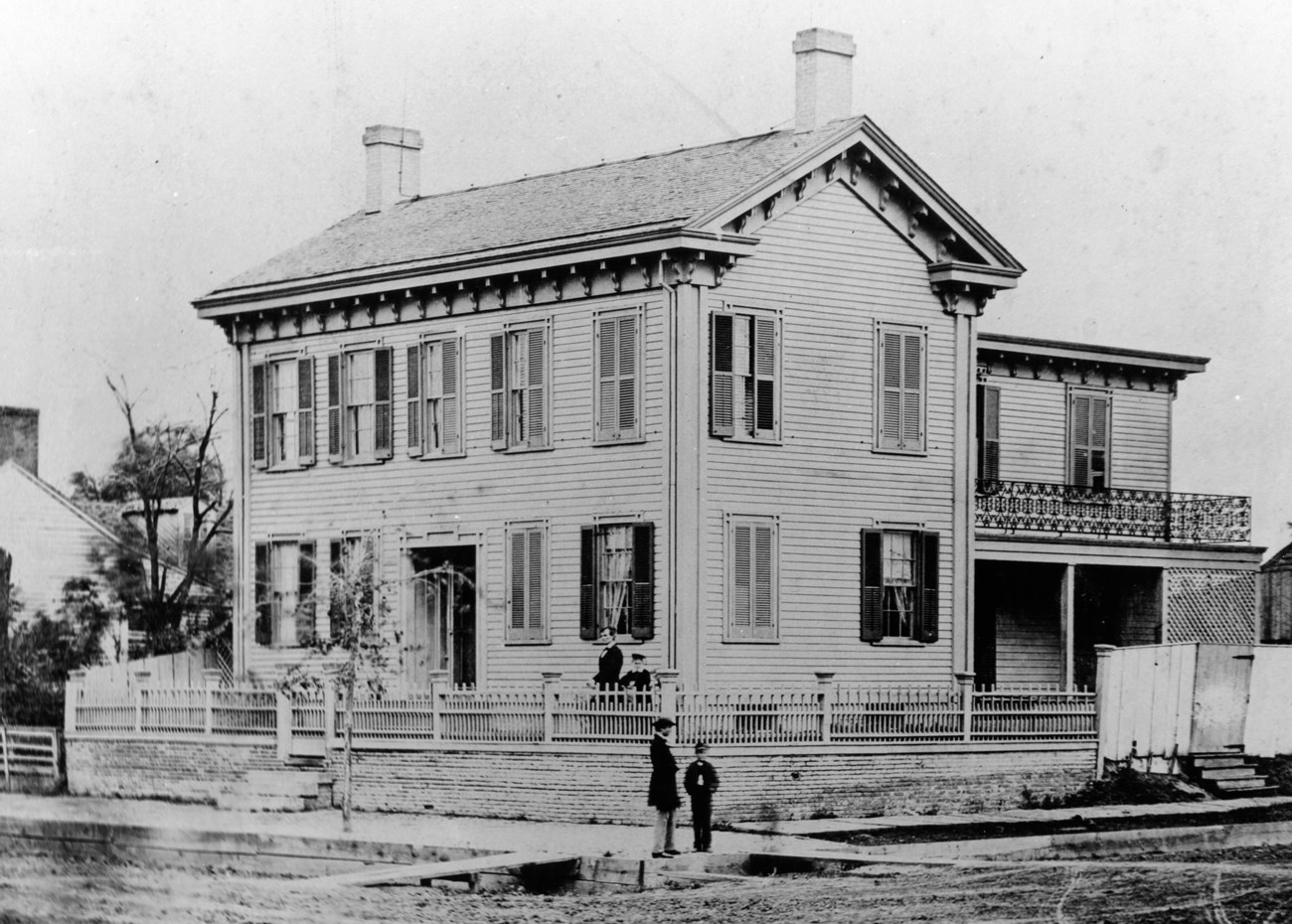 Black and white photo of the Lincoln Home, a 2 story wooden house on a brick retaining wall with a fence. Abraham Lincoln and his son stand behind front yard fence.