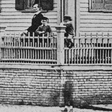 Close up image of Abraham Lincoln and his sons Willie and Tad standing in front yard behind fence, on front sidewalk a blurry shadow of a child is visible