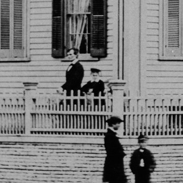 Close up image of Abraham Lincoln and his sons Willie and Tad standing in front yard behind fence. Tad is obscured by the corner fencepost.