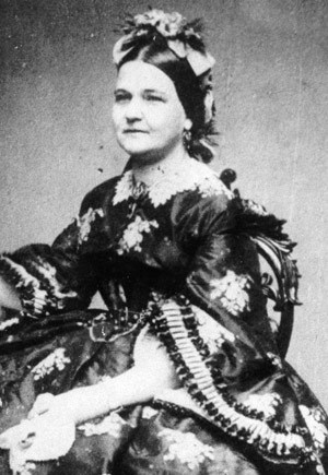 Mary Lincoln, a middle aged woman with dark hair parted in the middle and pinned back with a elaborate flowery headband. She wears a dark dress with long, flared sleeves and a white collar and pattern of flowers.