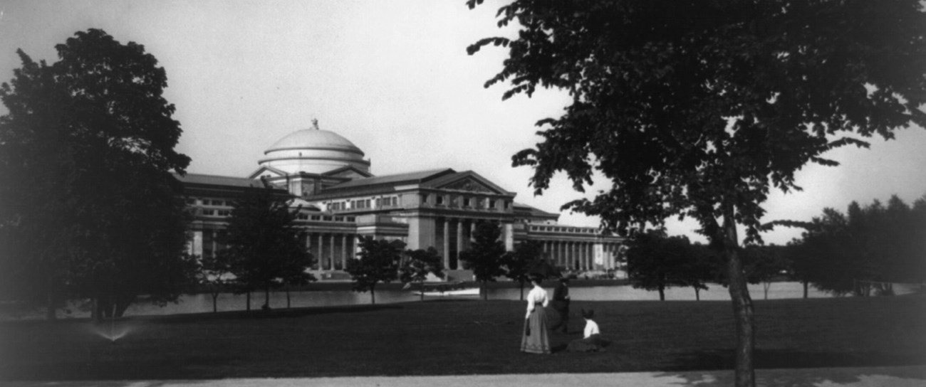 Black & White photo of a huge, long, grand building with pillars all along front side and a large domed roof in the center of the building