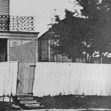 Close up photo of Lincoln Home back, south porch partially obscured by wooden latticework screen on porch, and a high white wooden fence around property. Shoddy gate door is built into fence. Dark, leafy tree peeks over the fence in backyard.