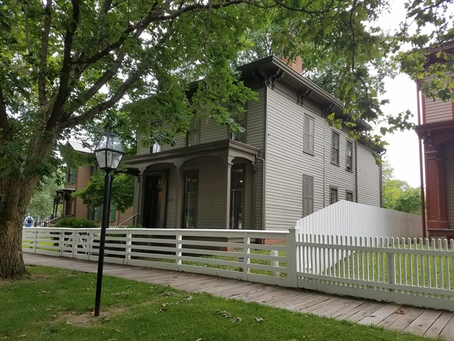 Gray, wooden 2-story House with darker gray trim, shutters, and front covered porch, shaded by strees