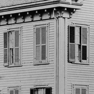 Close up photo of Lincoln Home shutters in corner of house, shutters are wood with two panels, one top and bottom, on each shutter. Shutters have ridges. One window has a shutter open, revealing blinds.